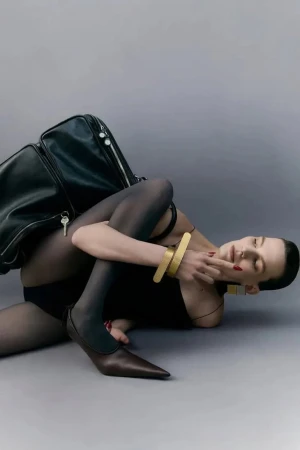 Model lying on grey studio floor wearing black bodysuit and dark tights next to a large black leather bag