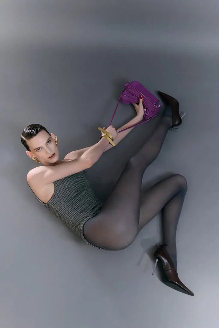 Model in grey patterned bodysuit holding a purple leather shoulder bag against a neutral grey studio backdrop