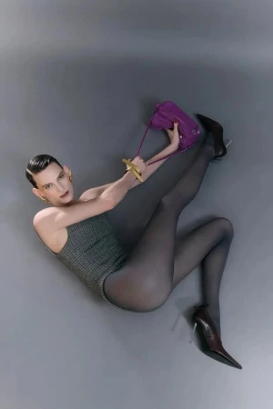 Model in grey patterned bodysuit holding a purple leather shoulder bag against a neutral grey studio backdrop