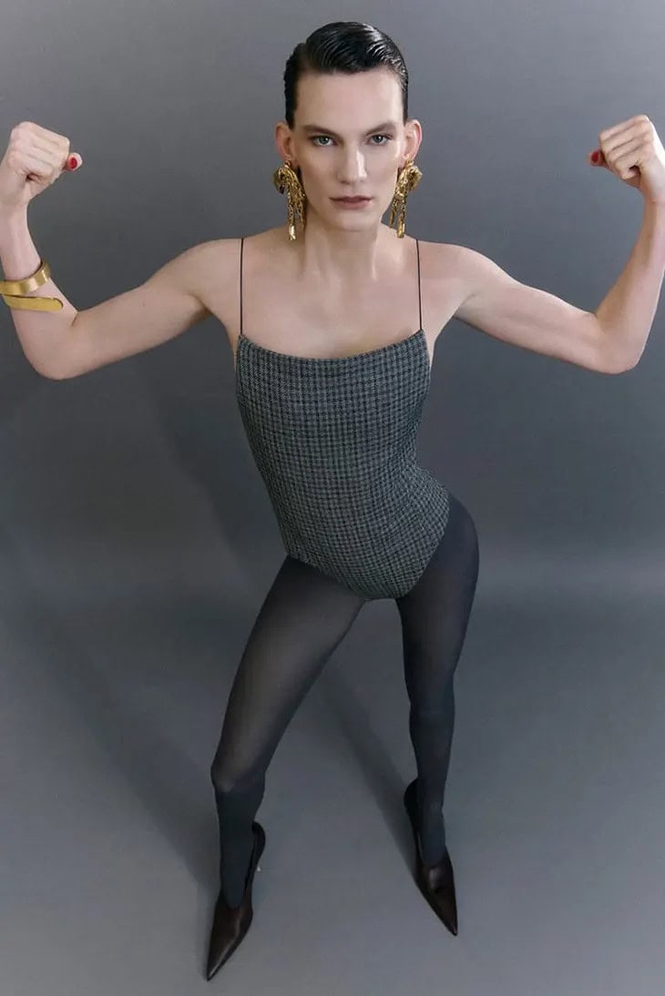 Model wearing a grey patterned bodysuit and dark tights flexing arms against a grey studio backdrop for Acne Studios.
