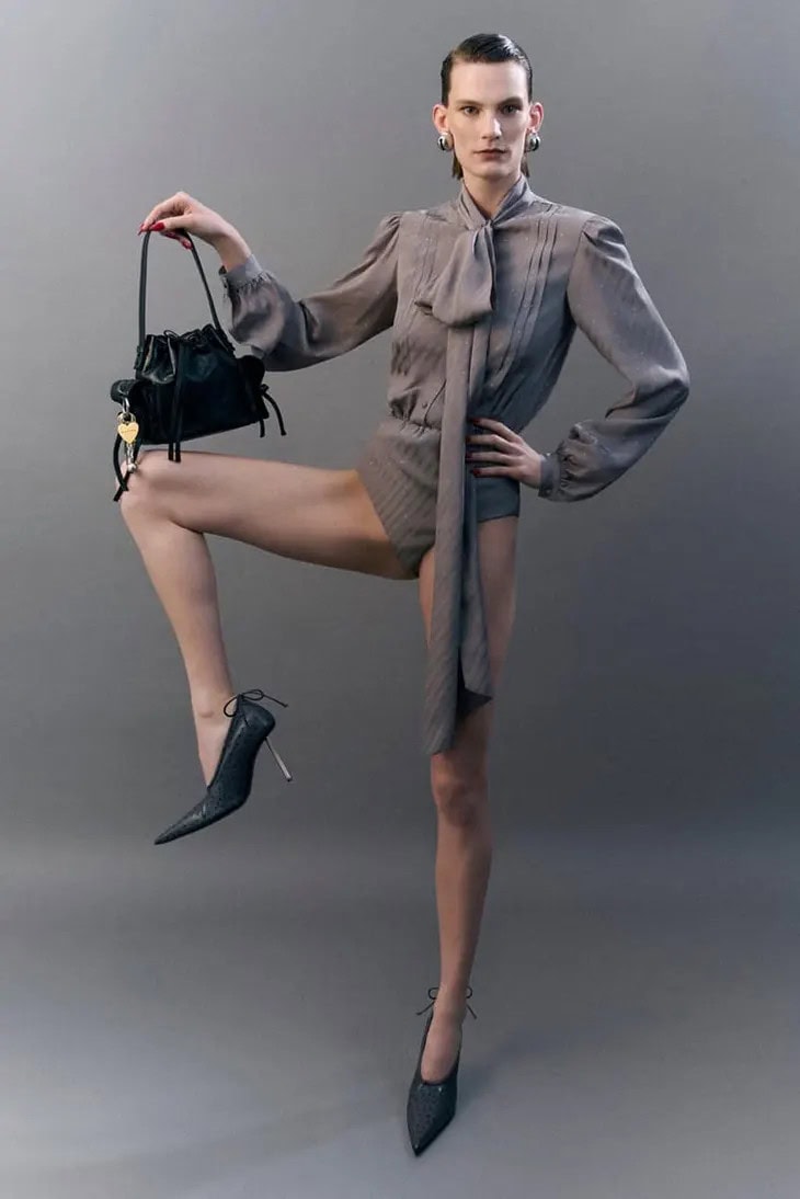 Model standing on one leg wearing a grey blouse and holding a black handbag against a grey background for Acne Studios.