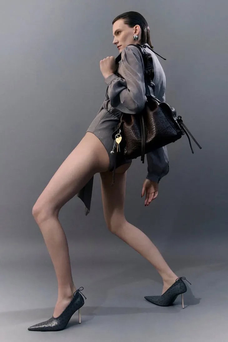 Model in grey leotard wearing black pumps carrying dark brown leather bag against grey background