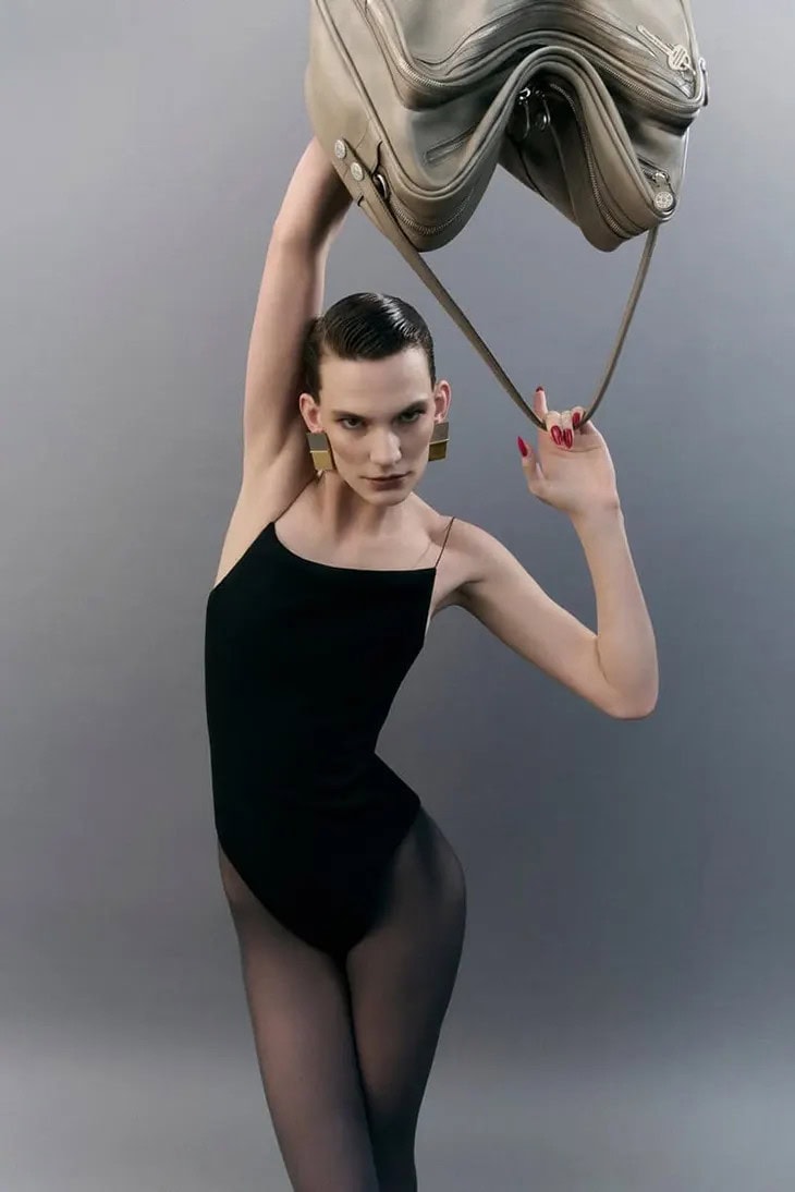 Model in black bodysuit holding a taupe leather shoulder bag overhead against a neutral grey studio background