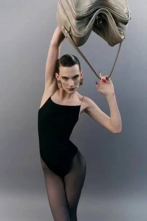 Model in black bodysuit holding a taupe leather shoulder bag overhead against a neutral grey studio background