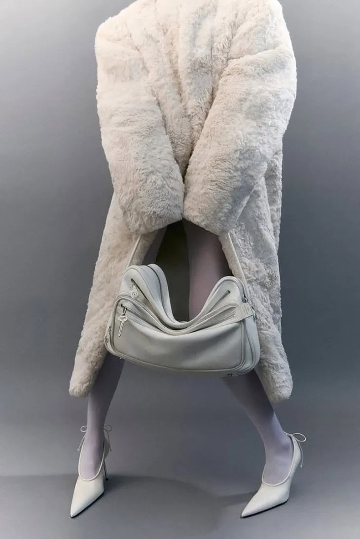 Model wearing a white faux fur coat and white heels holding an Acne Studios Camero bag against a grey seamless background