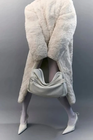 Model wearing a white faux fur coat and white heels holding an Acne Studios Camero bag against a grey seamless background