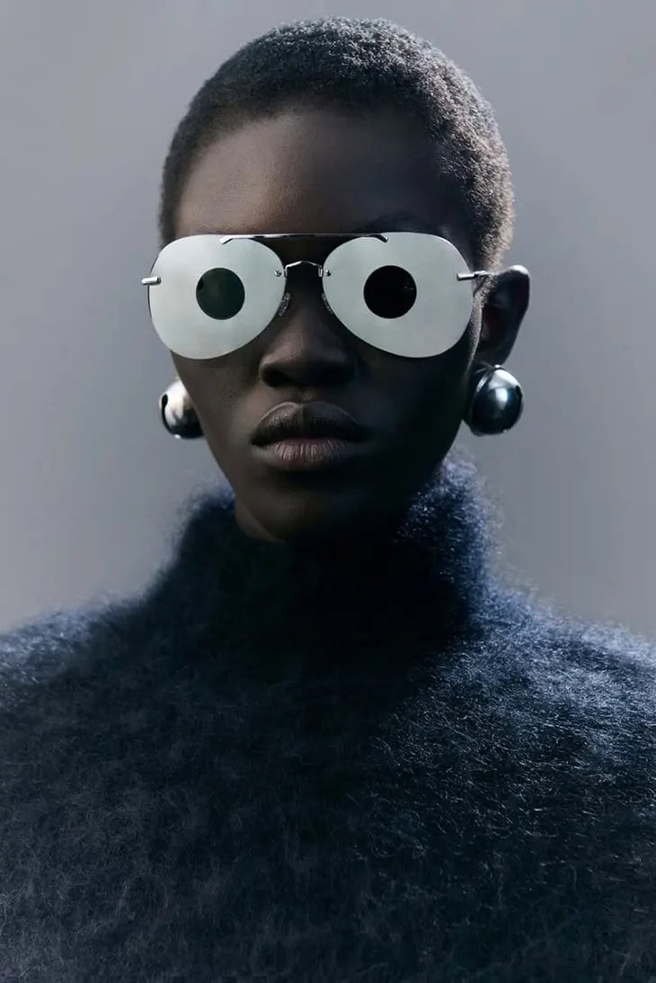 Close-up portrait of a model wearing large round white sunglasses with black pupils and oversized silver sphere earrings.