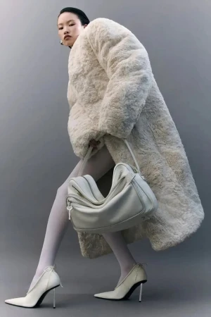 Model in fluffy cream coat holding a white shoulder bag against a grey seamless studio backdrop
