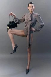 Model standing on one leg wearing a grey blouse and holding a black handbag against a grey background for Acne Studios.