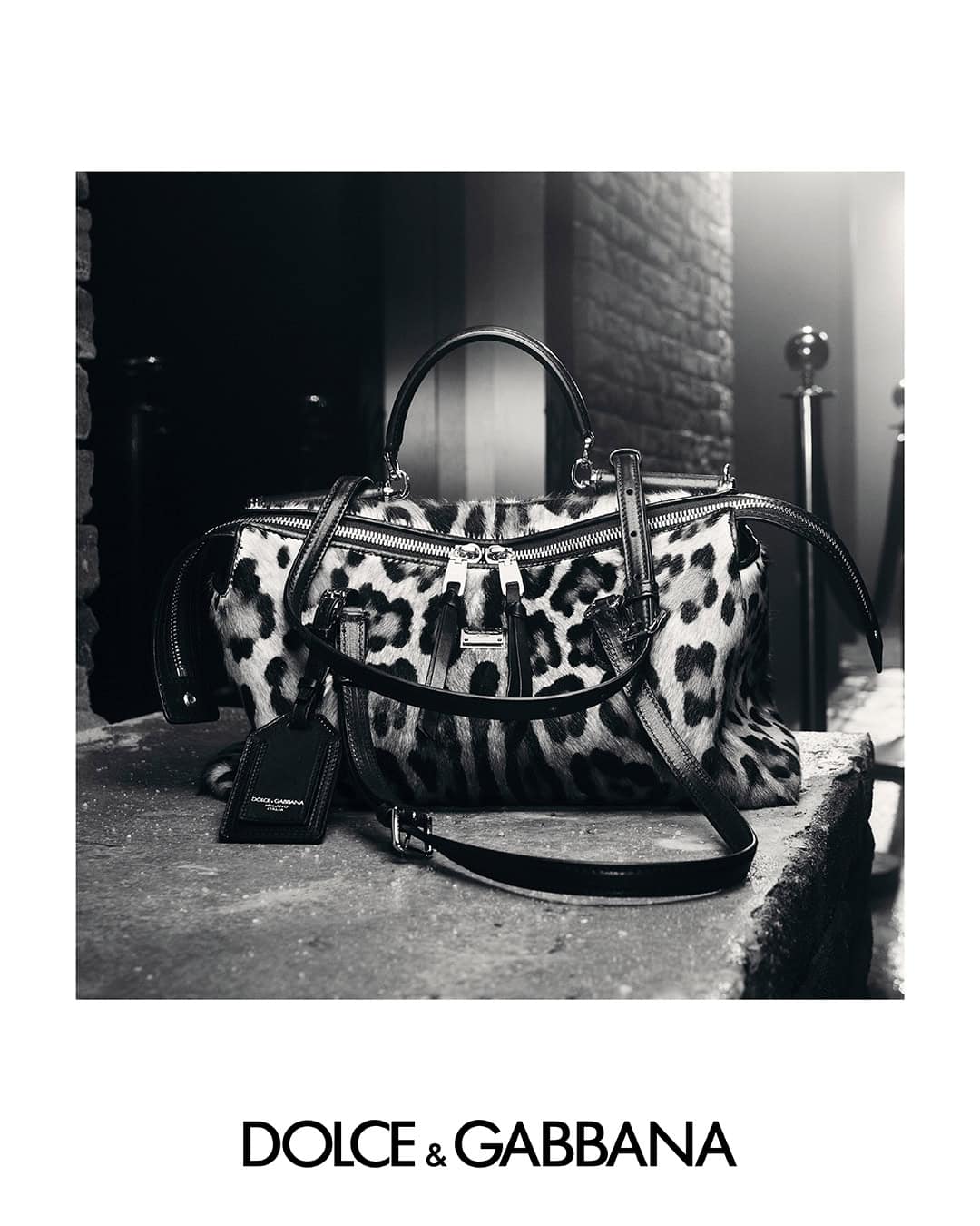 Black and white product shot of a leopard print handbag with top handles and a luggage tag resting on stone surface