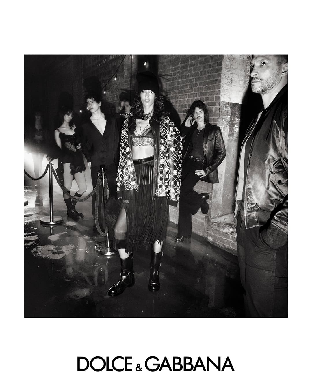 Black and white fashion campaign image featuring models in dark attire standing on a wet surface against a brick wall