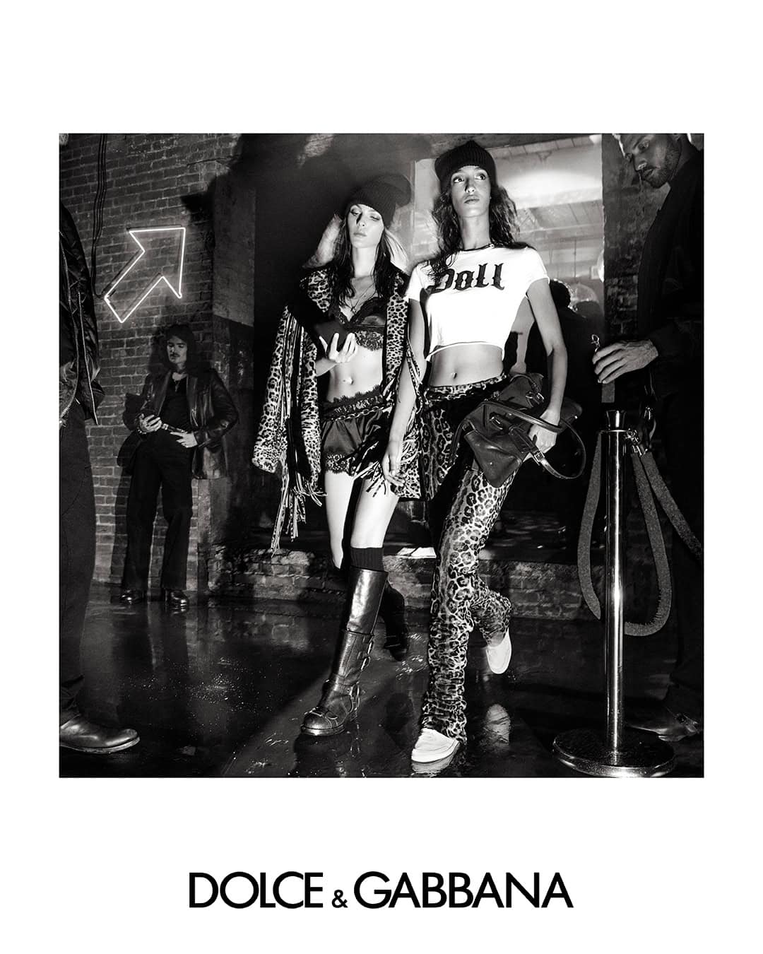 Black and white fashion campaign image featuring two female models wearing beanies and crop tops against a brick wall backdrop.