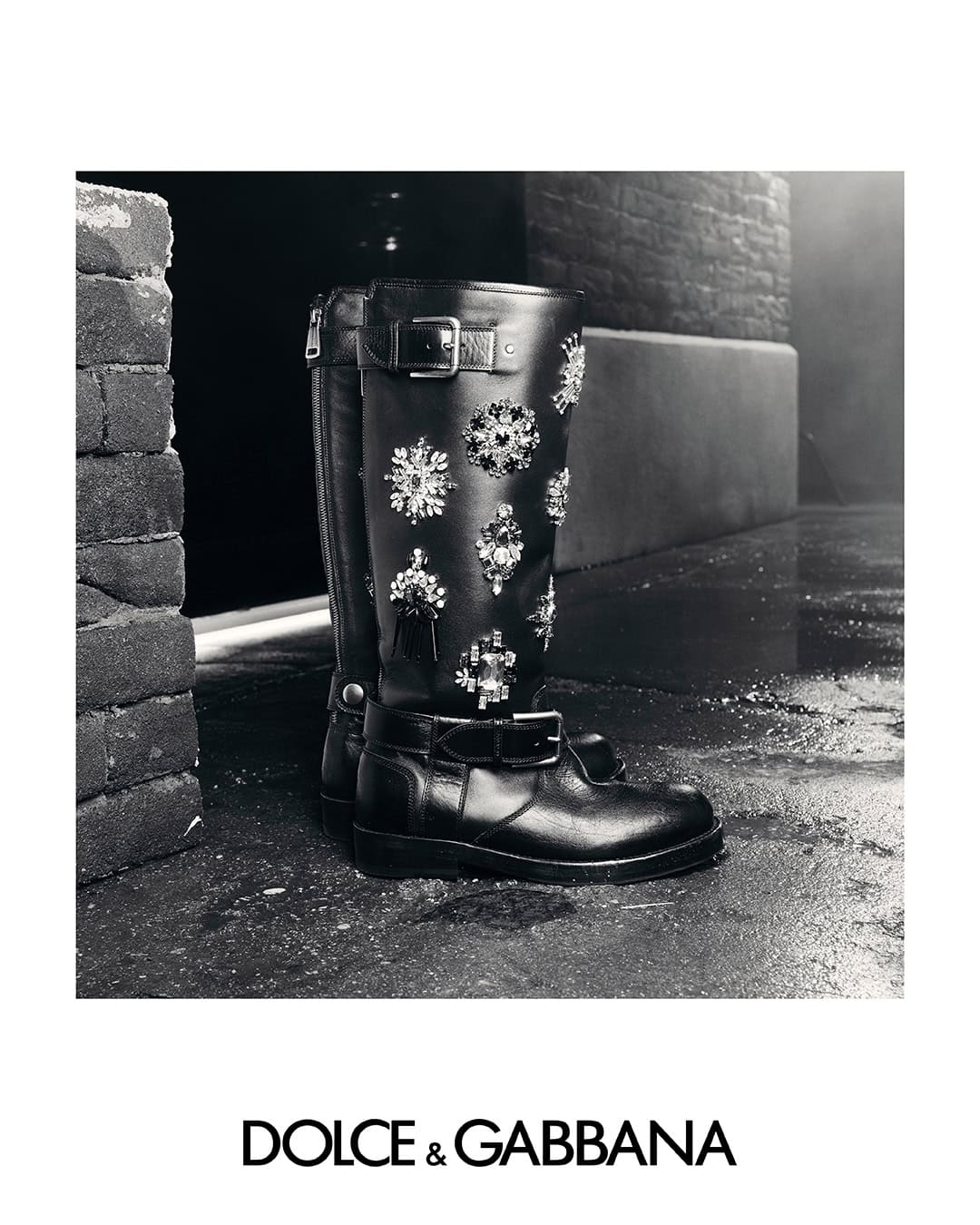 Black embellished leather boots with crystal details on a wet concrete surface for Dolce & Gabbana Fall 2025 campaign.