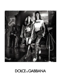 Black and white fashion campaign image featuring two female models wearing beanies and crop tops against a brick wall backdrop.