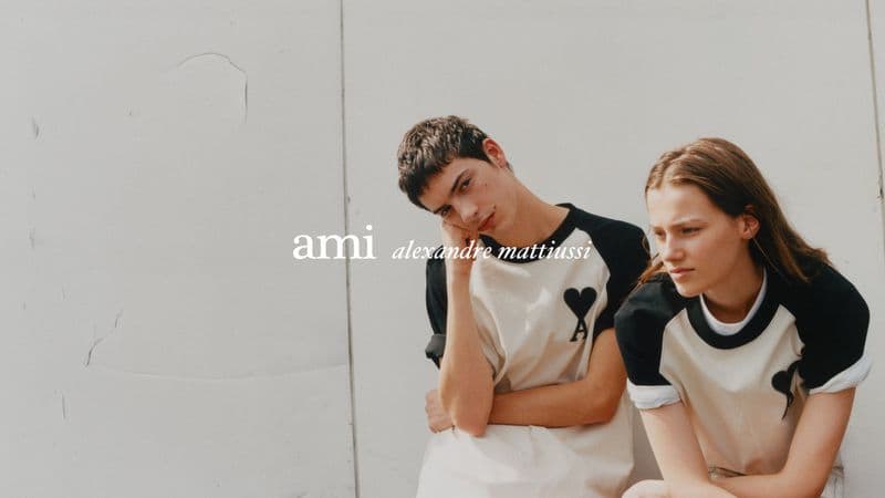 Two models wearing white raglan t shirts with black sleeves and heart logos posed against a neutral concrete wall