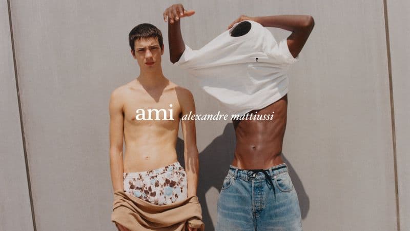 Two shirtless male models standing against a neutral concrete wall for an Ami ad campaign, one pulling off a white t-shirt.