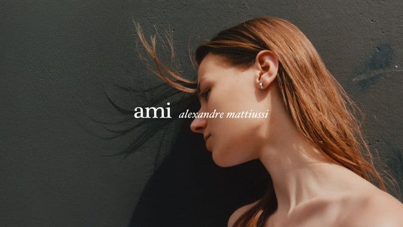 Profile portrait of a woman with long hair blowing in the wind against a dark textured wall for an Ami campaign.