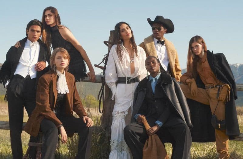 Group of six models in western-inspired formalwear posing outdoors by a wooden fence with distant mountains.