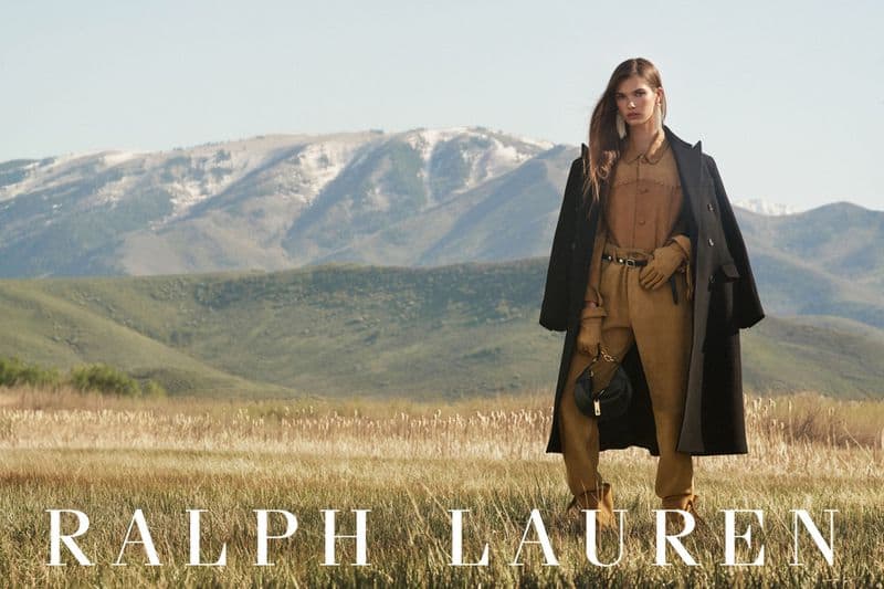 Model standing in a grassy field wearing a brown suit and long black coat with mountain backdrop for Ralph Lauren