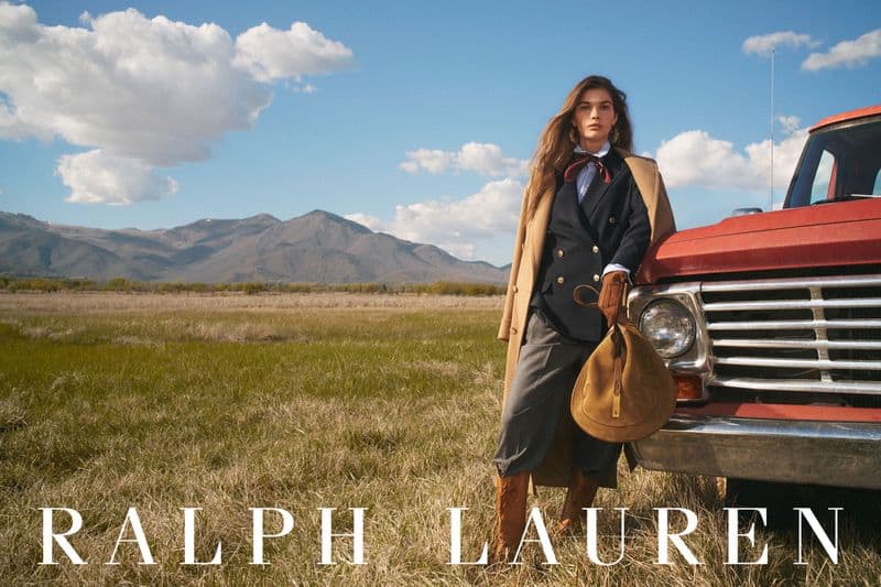 Model standing in a grassy field beside a red truck wearing a blazer and holding a brown bag against mountains and a blue sky