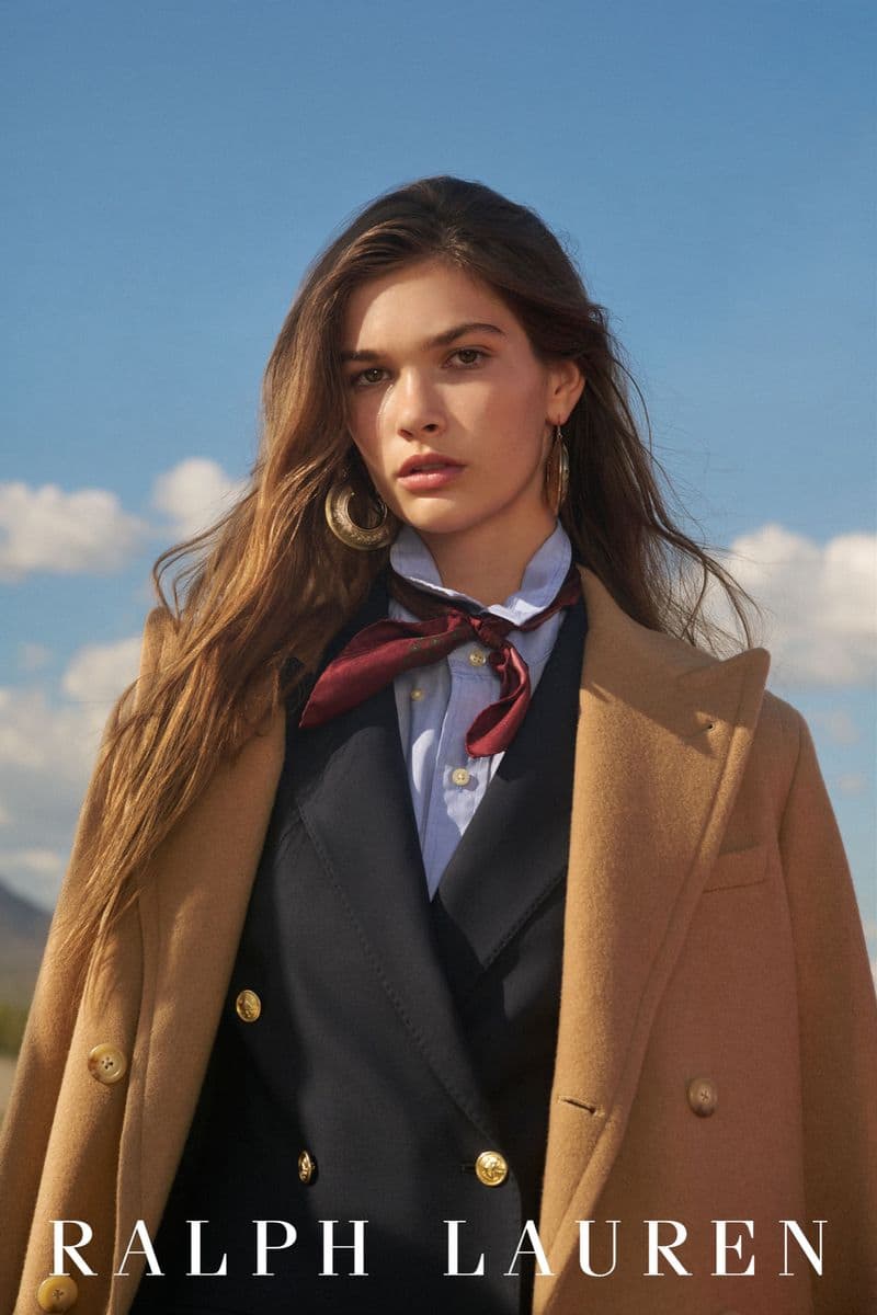 Portrait of a female model wearing a camel coat, dark blazer, light shirt, and burgundy scarf against a blue sky