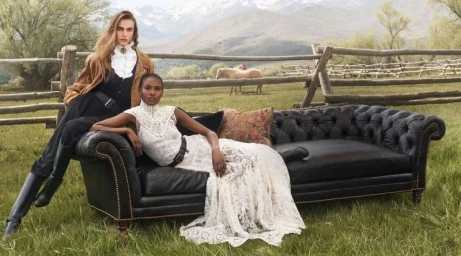 Two female models posing outdoors on a black leather sofa with a rustic wooden fence and mountain landscape background.