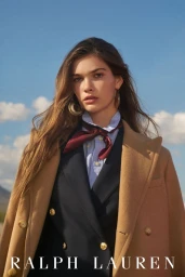 Portrait of a female model wearing a camel coat, dark blazer, light shirt, and burgundy scarf against a blue sky