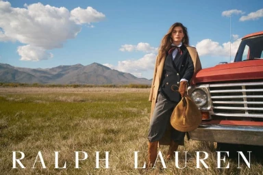 Model standing in a grassy field beside a red truck wearing a blazer and holding a brown bag against mountains and a blue sky
