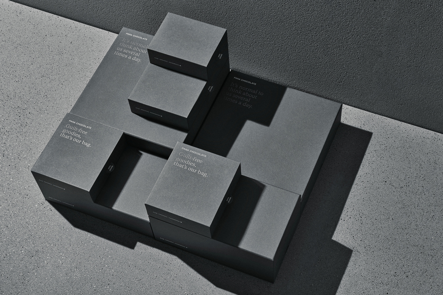 Overhead view of dark grey Pana Chocolate packaging boxes arranged symmetrically on a textured concrete surface with sharp shadows.