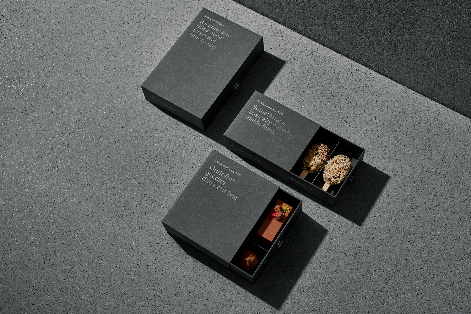 Three dark grey Pana Chocolate boxes arranged on a textured grey concrete surface, with one box open showing chocolates.