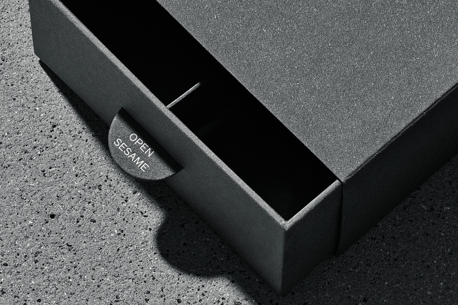 Close-up of a dark grey textured cardboard drawer box with open sesame text on the pull tab resting on a speckled concrete surface.