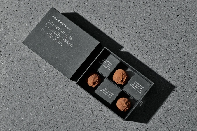 An open dark grey Pana Chocolate gift box containing chocolate truffles resting on a textured concrete surface.