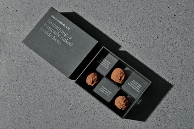 An open dark grey Pana Chocolate gift box containing chocolate truffles resting on a textured concrete surface.