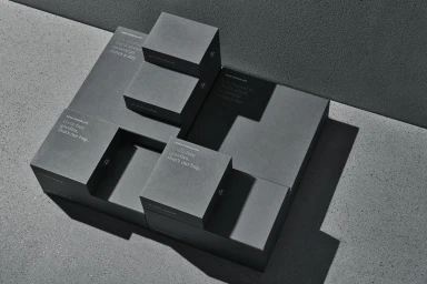 Overhead view of dark grey Pana Chocolate packaging boxes arranged symmetrically on a textured concrete surface with sharp shadows.