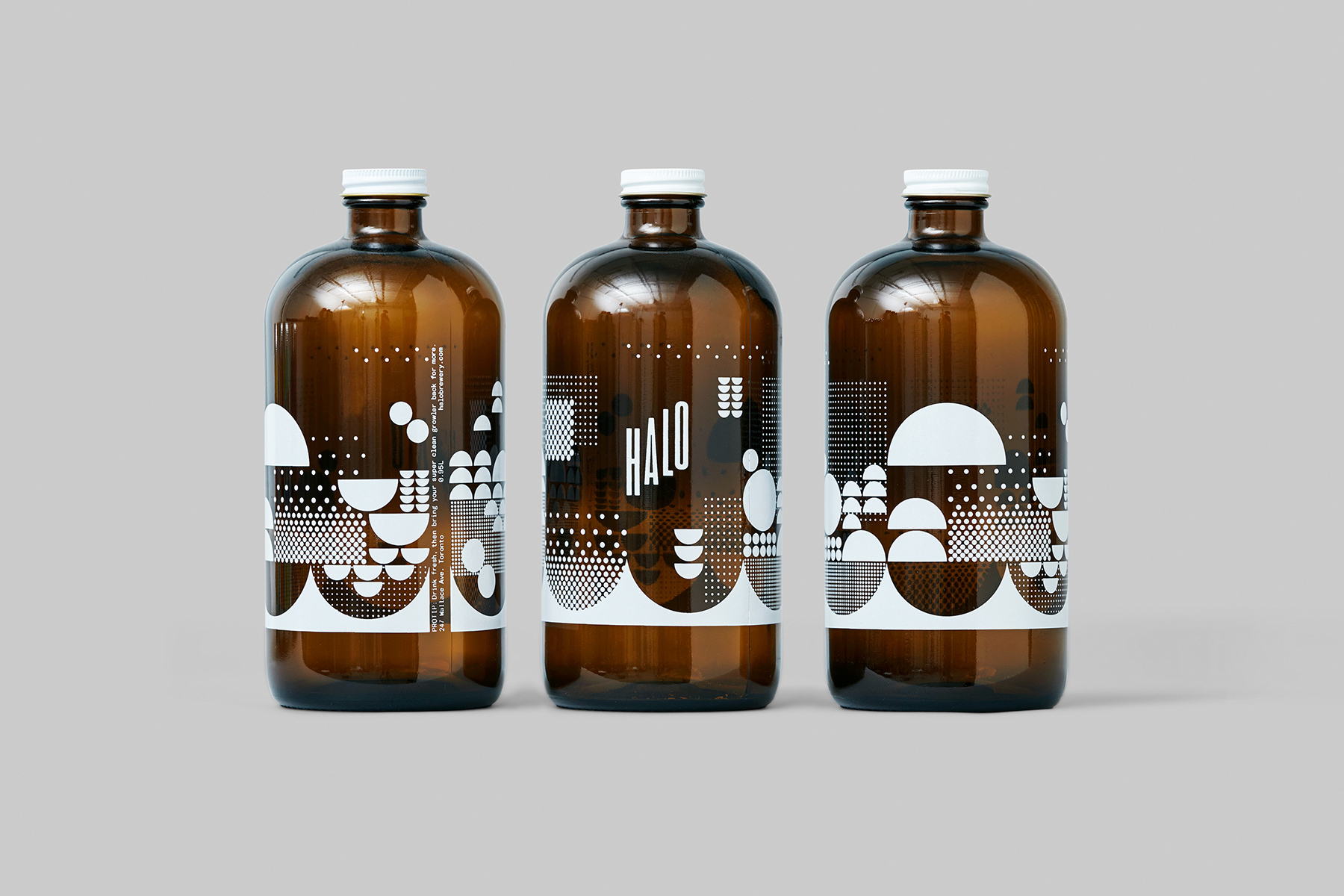 Three amber glass beer bottles with white geometric labels and caps against a grey studio background.