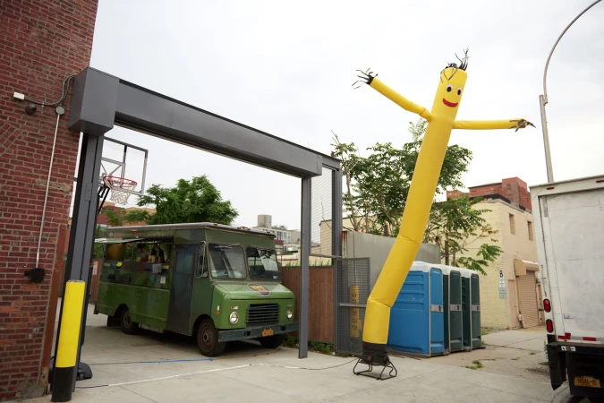 Bright yellow inflatable tube man standing beside a green food truck in an alleyway with brick buildings.