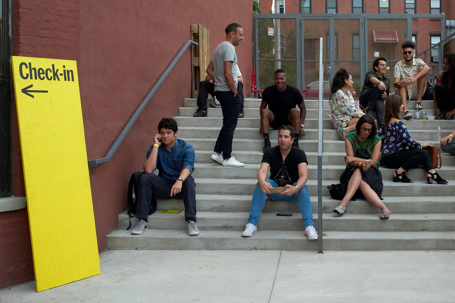 People gathered on outdoor concrete steps near a tall yellow Check-in sign on a red brick building wall