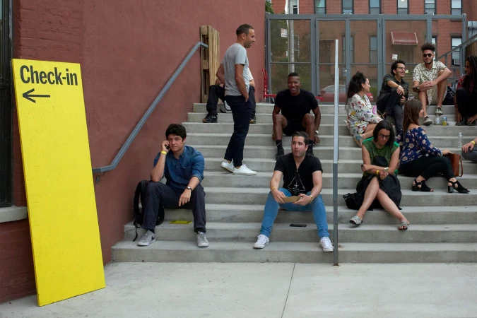 People gathered on outdoor concrete steps near a tall yellow Check-in sign on a red brick building wall