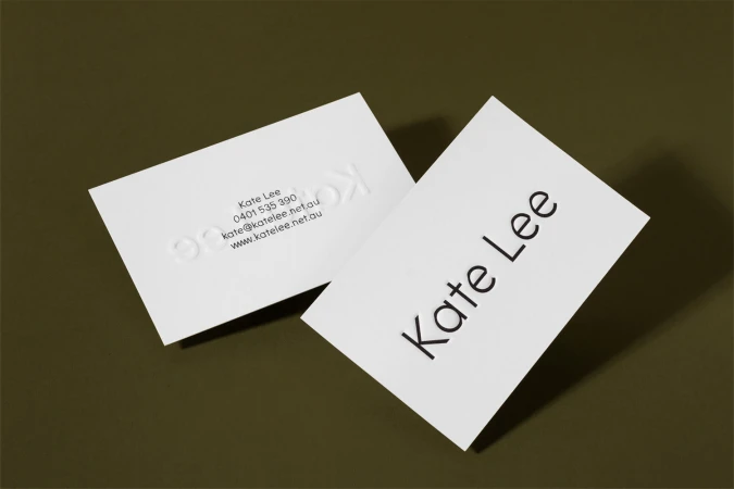 Two white business cards with debossed and printed typography resting on a dark olive green surface.