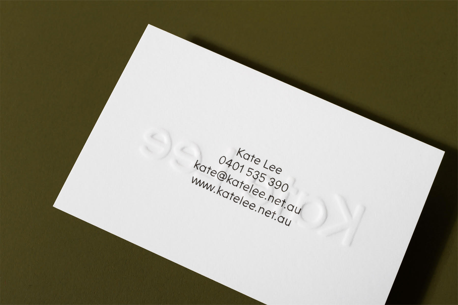 White business card with embossed typography and contact details resting on an olive green surface.
