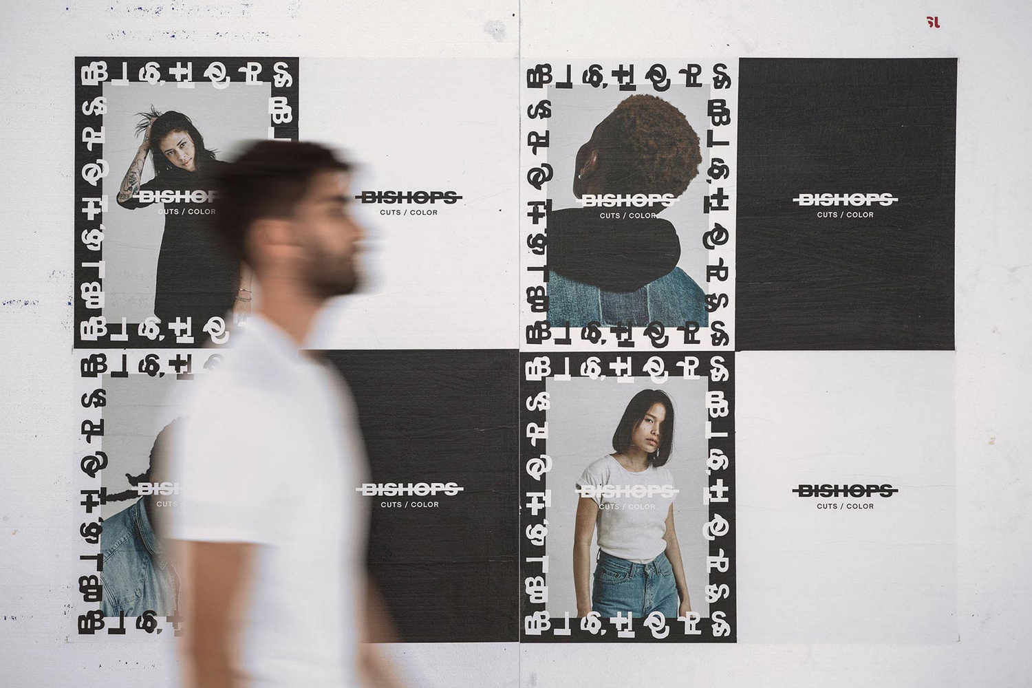 A man in a white t-shirt walks past a wall displaying black and white Bishops brand identity posters and portraits.