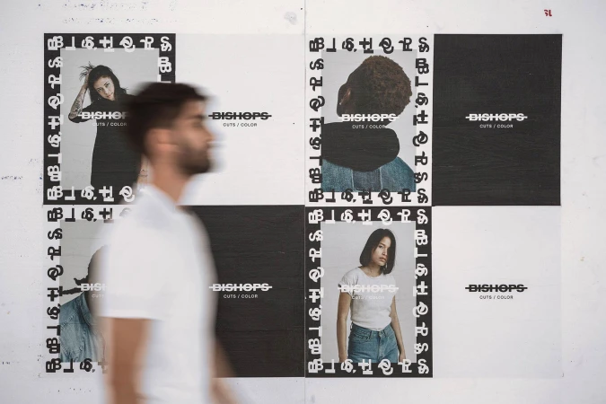 A man in a white t-shirt walks past a wall displaying black and white Bishops brand identity posters and portraits.