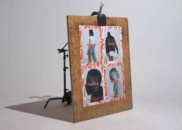 A large wooden board displaying a four-panel poster with red typographic branding is propped up in a bright studio space.