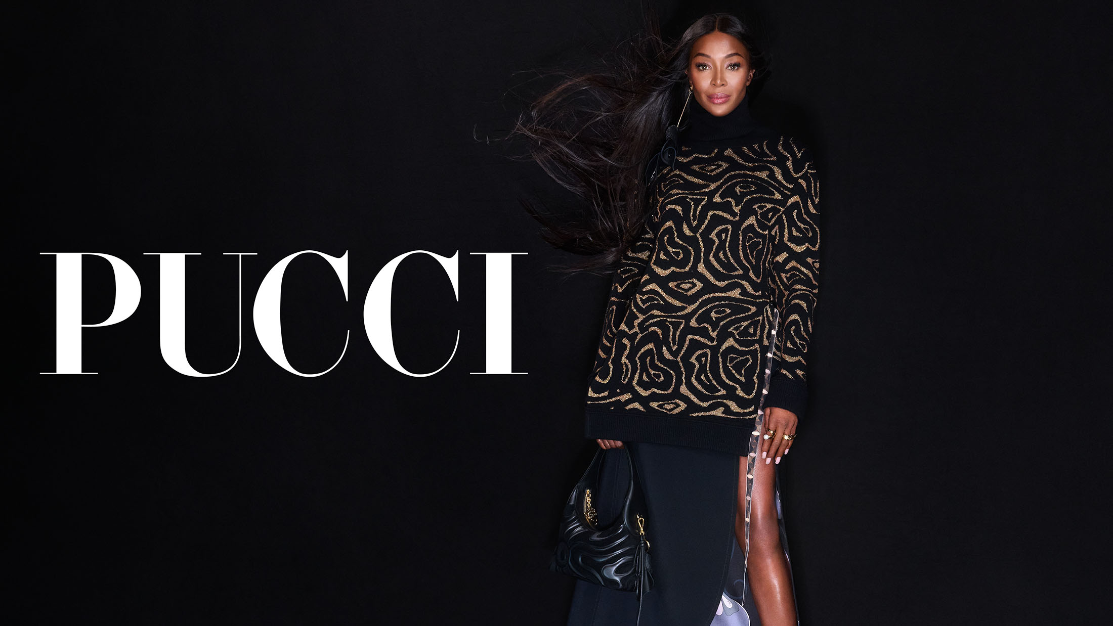 Naomi Campbell wearing a patterned black and gold turtleneck sweater and holding a black handbag against a black background for Pucci