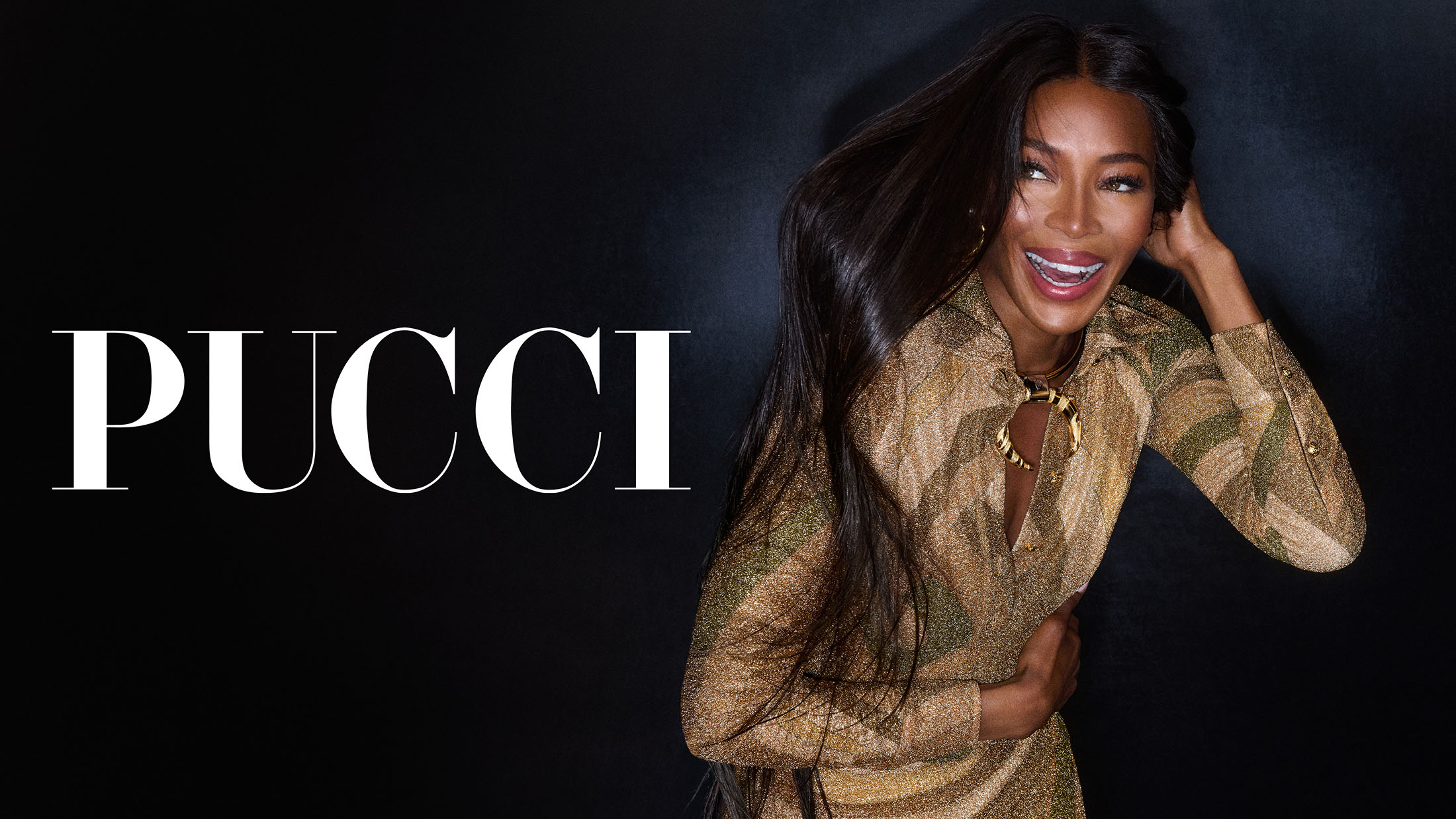 Naomi Campbell smiling wearing a metallic patterned long-sleeve top against a dark background with PUCCI typography