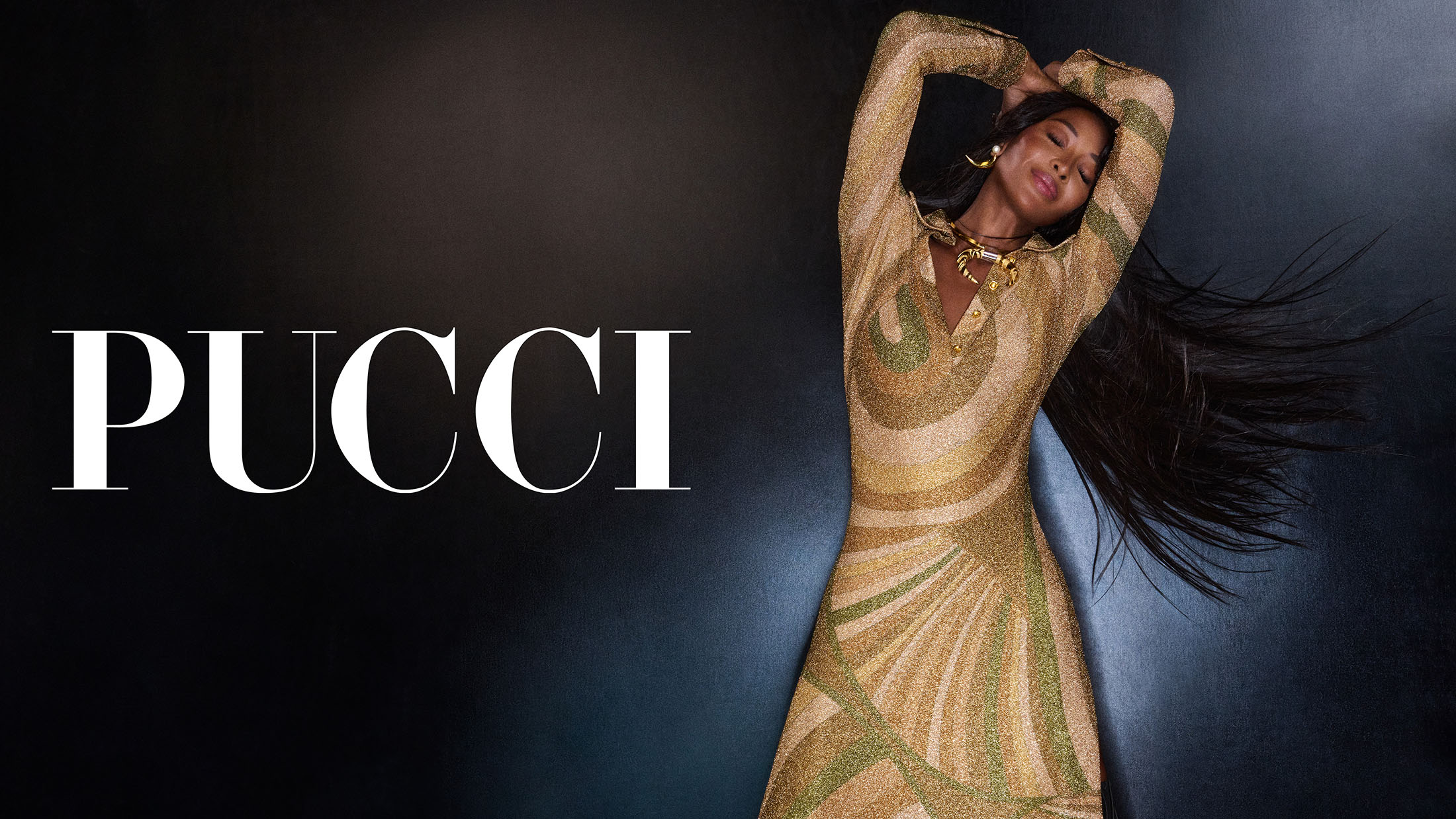Naomi Campbell modeling a patterned gold and green Pucci dress with long dark hair against a dark studio background