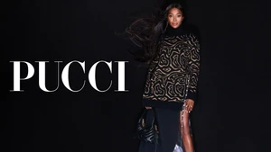 Naomi Campbell wearing a patterned black and gold turtleneck sweater and holding a black handbag against a black background for Pucci