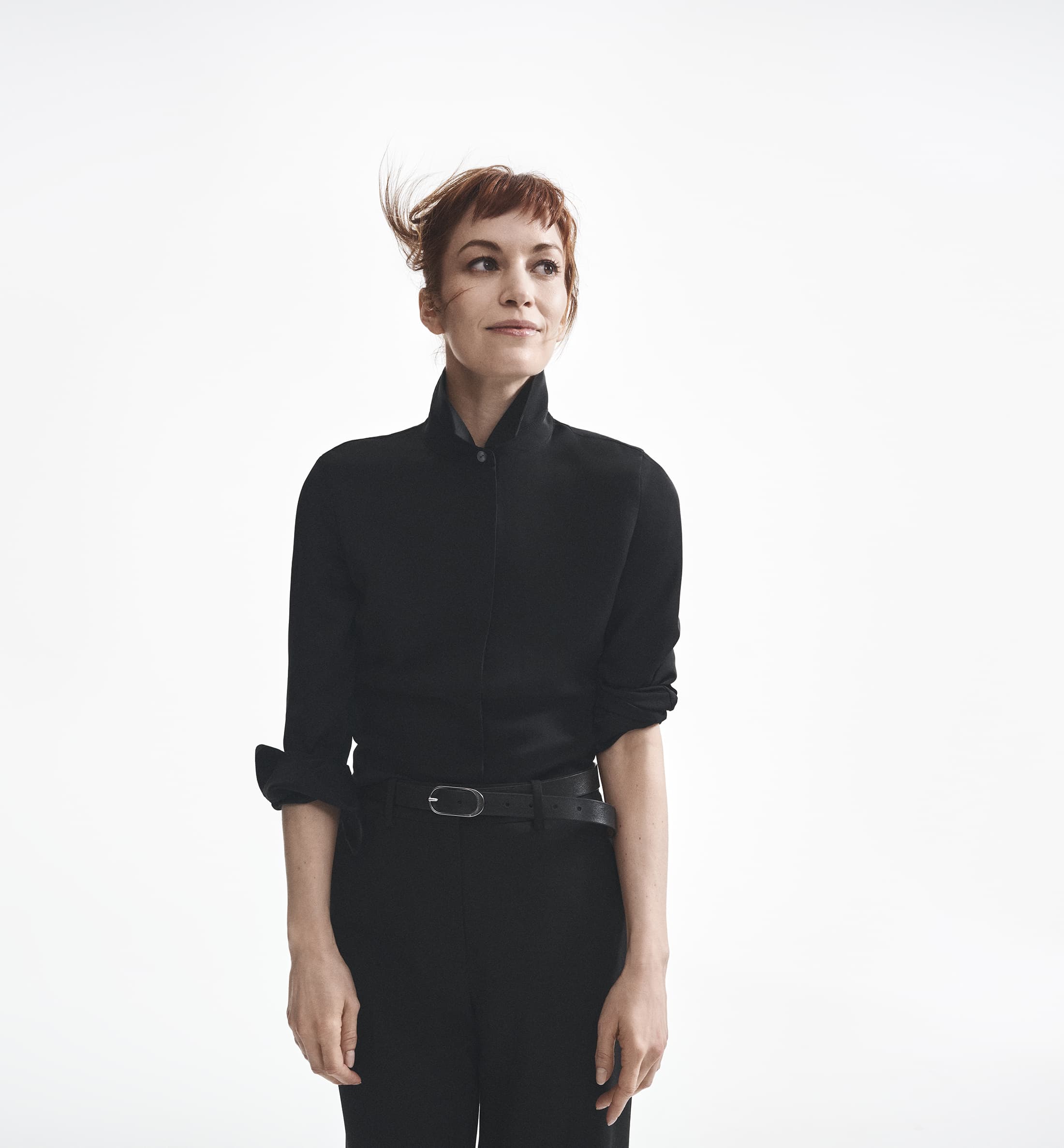 Britt Lower wearing a black button-down shirt and trousers against a bright white background