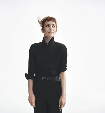 Britt Lower wearing a black button-down shirt and trousers against a bright white background