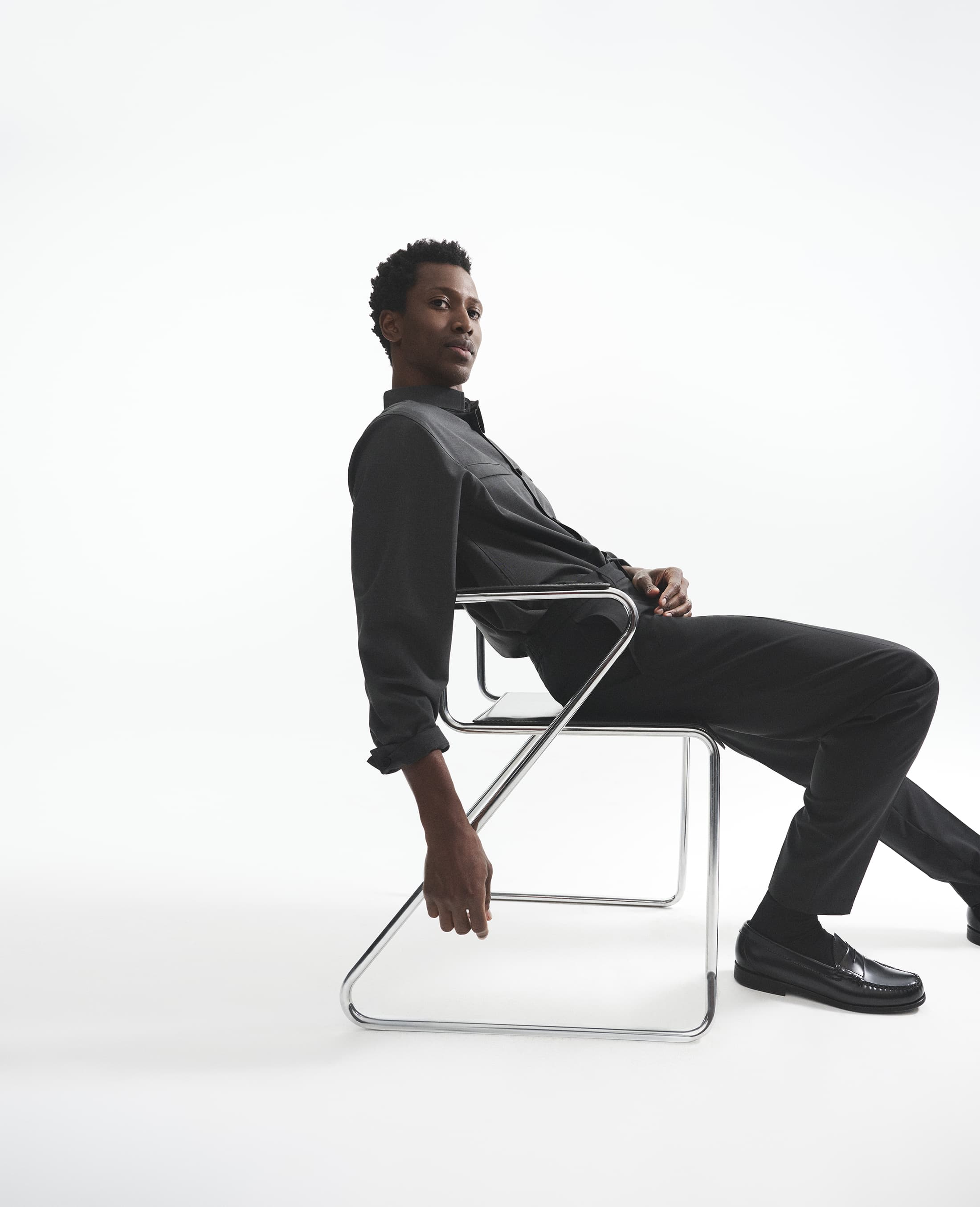 Man seated on a modern chrome cantilever chair against a plain white studio background wearing dark shirt and trousers.